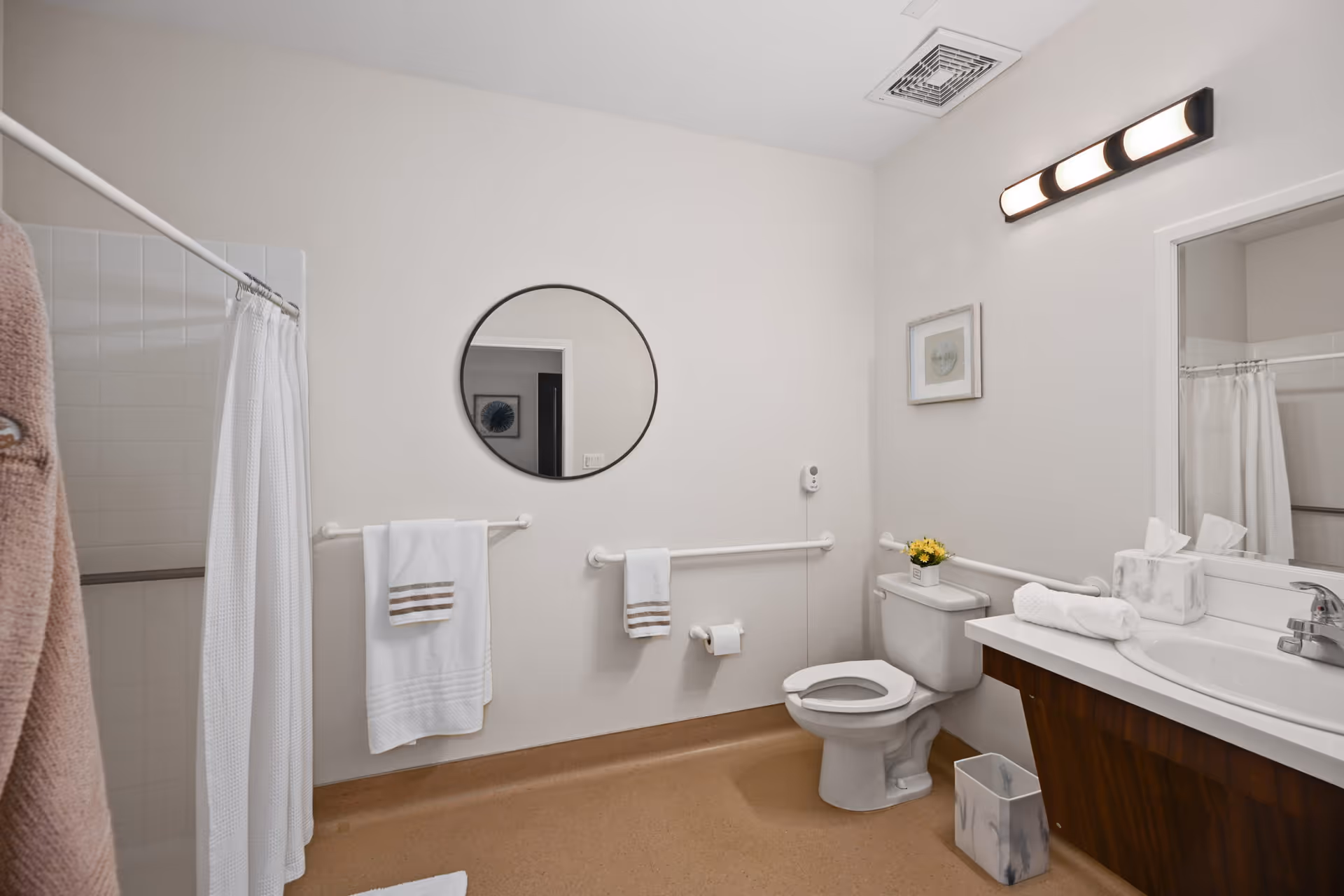 A clean and accessible bathroom with beige flooring and white walls. The room features a shower with a white curtain, a round mirror above a towel rack with white towels, a toilet with a small flower pot on the tank, and a sink with a marble tissue box and a rolled white towel. There are safety grab bars installed near the toilet and along the wall. A rectangular light fixture with four bulbs is mounted above the sink mirror.