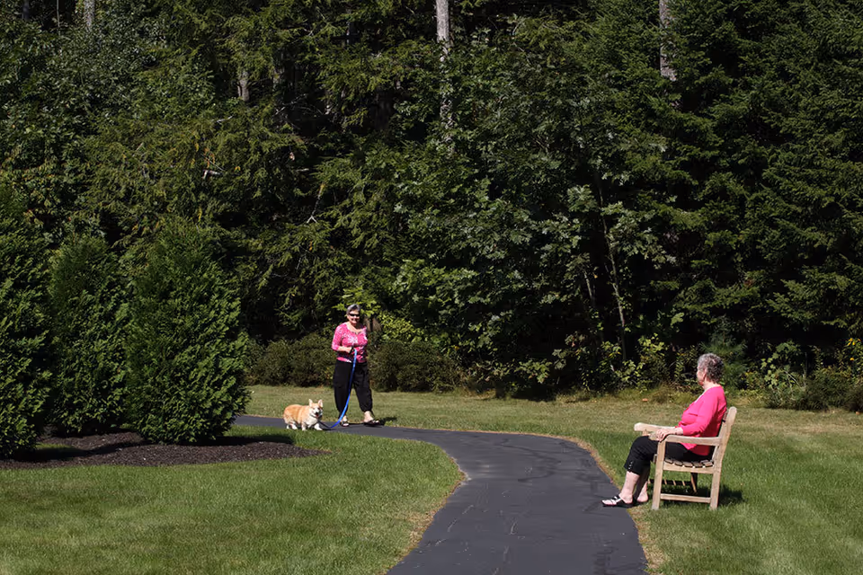 Two women in a grassy outdoor area — one walking a corgi on a paved path and another seated on a bench near trees.