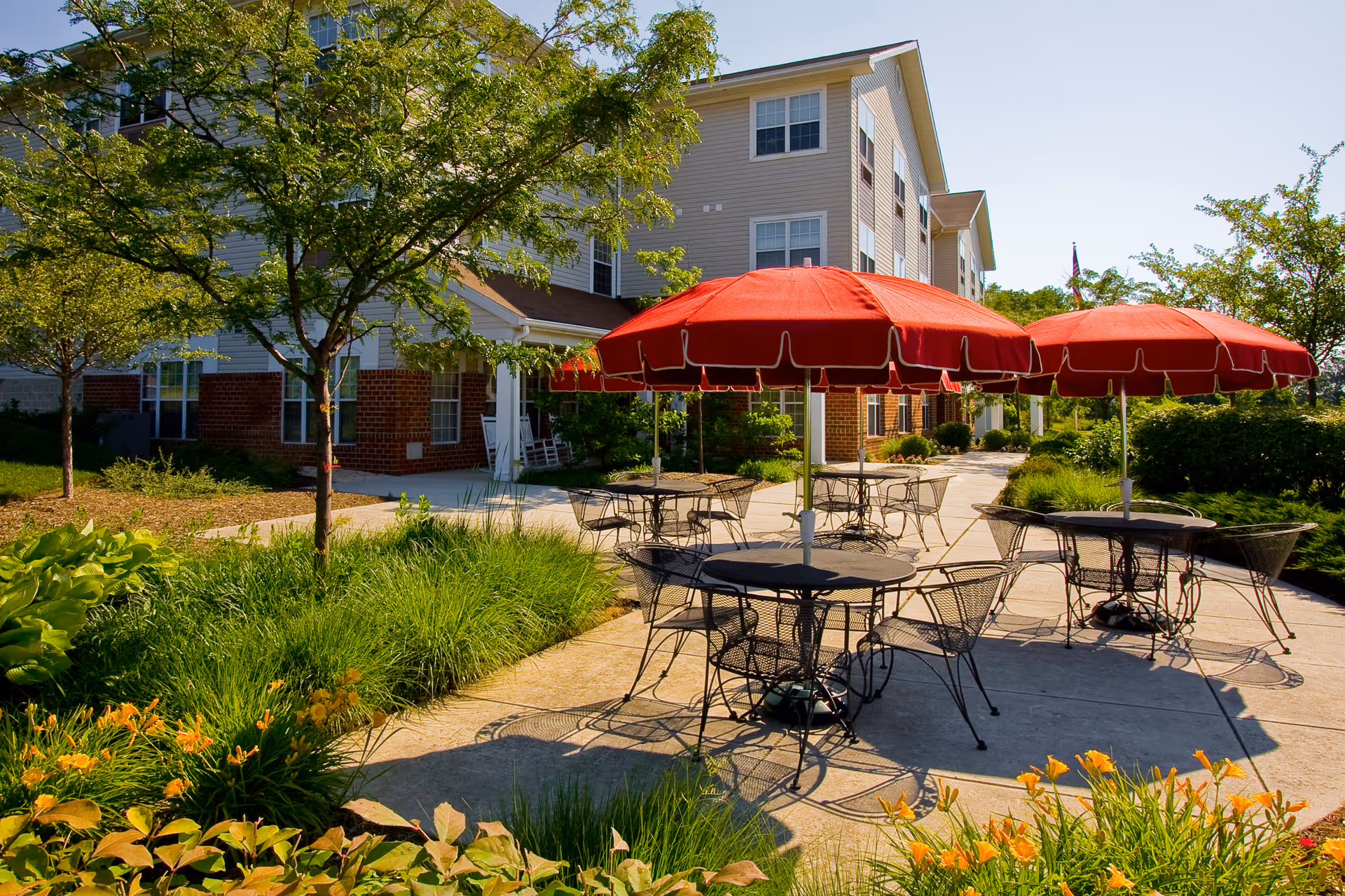 Outdoor patio area at a senior living facility with several round metal tables and chairs under red umbrellas, surrounded by green plants and trees, with a multi-story building in the background under a clear blue sky.