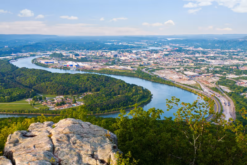 A panoramic aerial view of a river winding through a green forested area with a cityscape in the background under a partly cloudy sky.