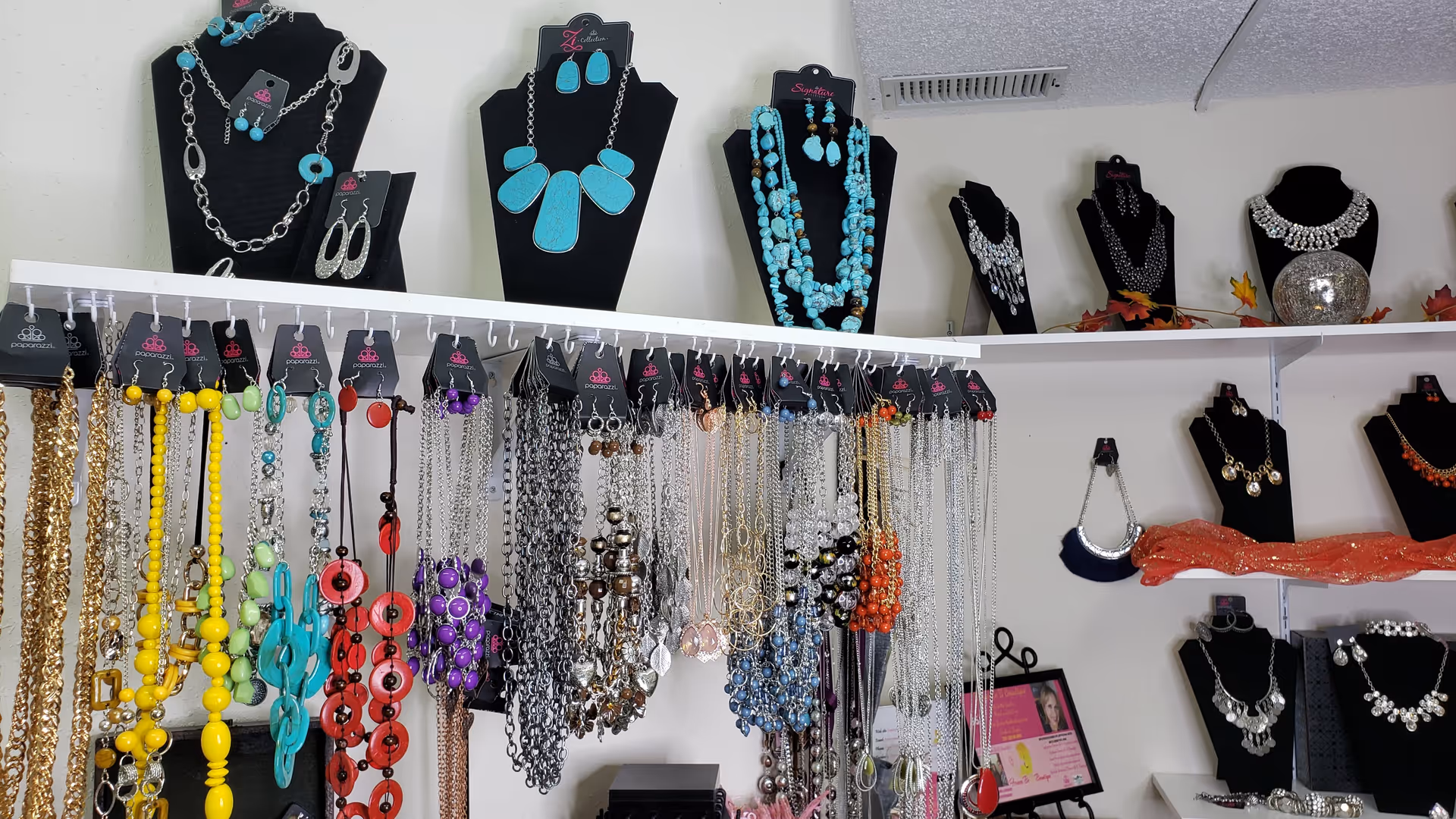 Display of various colorful necklaces and earrings hanging on hooks and displayed on black necklace stands against a white wall.