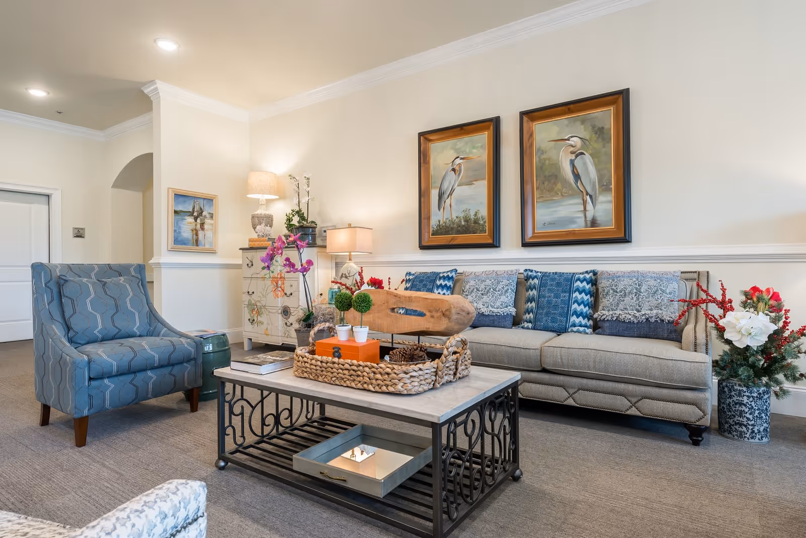 A cozy living room with a beige sofa adorned with blue and patterned throw pillows, a blue patterned armchair, and a coffee table with decorative items including a woven basket, small plants, and books. The walls are light-colored with two framed paintings of birds hanging above the sofa. There are two table lamps and a floral arrangement in a blue and white vase on the floor.