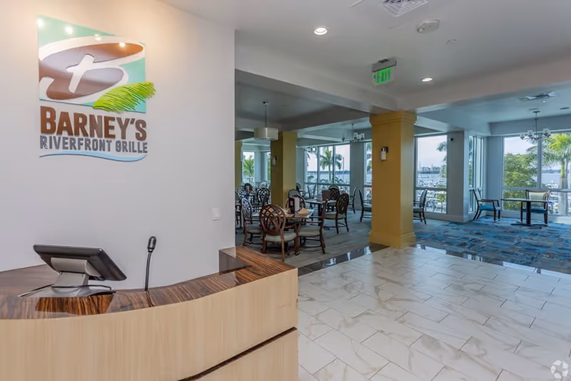 Reception desk area with a sign reading 'Barney's Riverfront Grille' on the wall. Beyond the desk is a dining area with tables and chairs, large windows showing a view of palm trees and water outside.