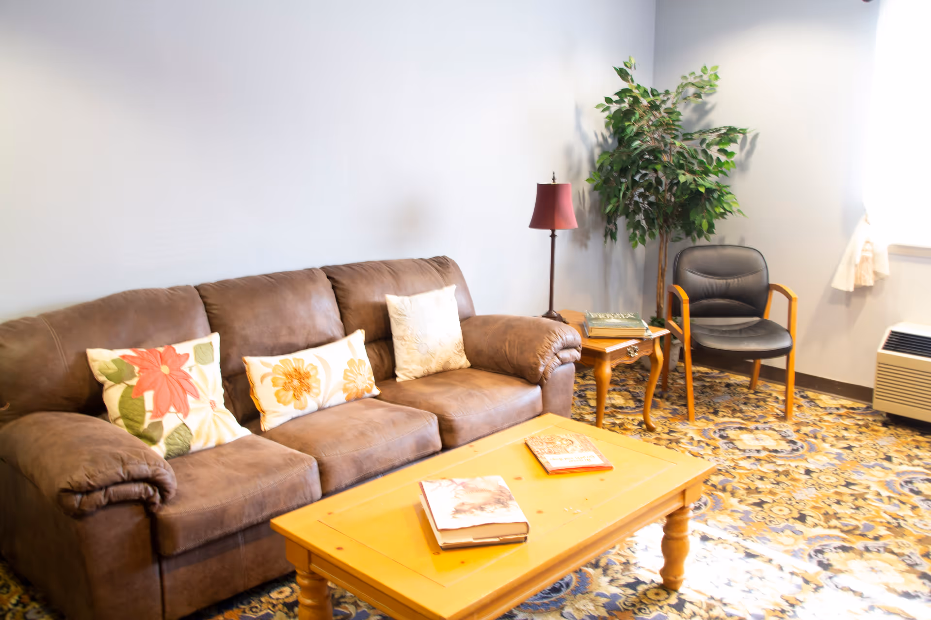A cozy living room with a brown sofa adorned with three floral-patterned pillows, a wooden coffee table with two books on it, a side table with a lamp and books, a black chair with wooden armrests, a potted plant, and a window with white curtains letting in natural light. The floor is covered with a patterned carpet.