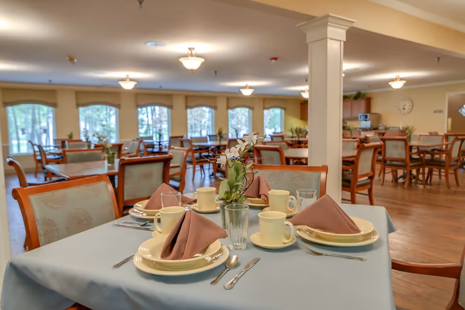 A bright dining room with a table in the foreground set with plates, cups, folded napkins, and a floral centerpiece, with more tables and chairs in the background.