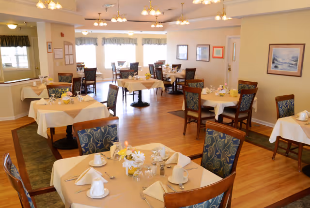 A bright and spacious dining room with multiple tables covered in beige tablecloths, each set with white cups, napkins, silverware, and small floral centerpieces. The room has wooden floors, several chairs with patterned upholstery, large windows with valances letting in natural light, and framed artwork on the walls.