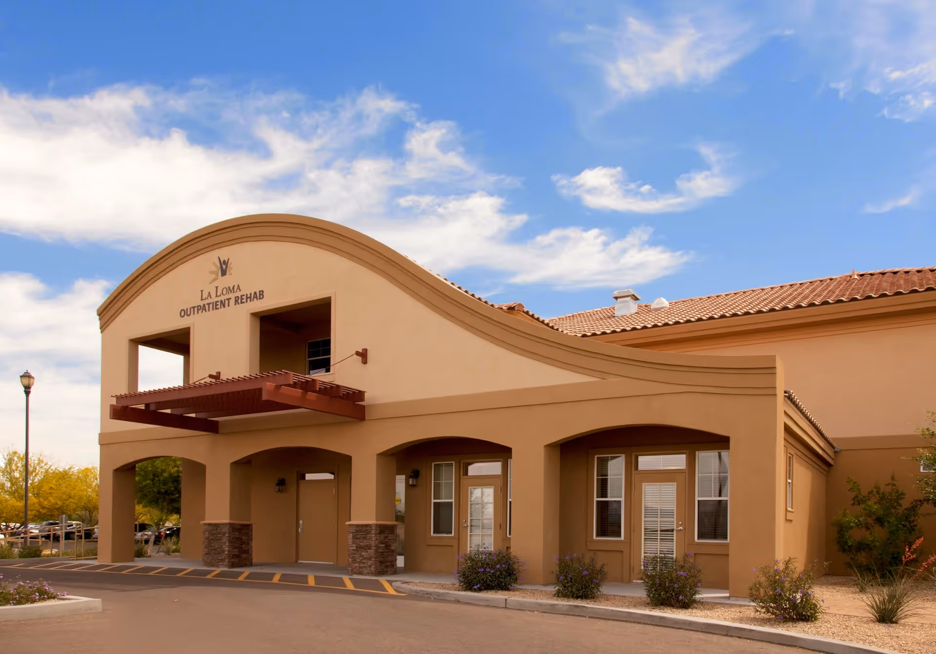 Exterior view of La Loma Outpatient Rehab building with a curved roofline, beige stucco walls, and a red tile roof under a partly cloudy blue sky. There are small bushes and a parking area in front.