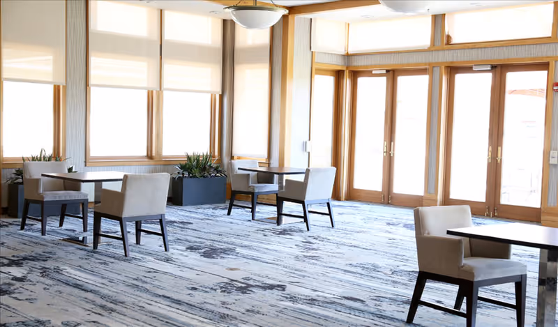 Bright common dining area with small tables and upholstered chairs beside large windows and glass doors.