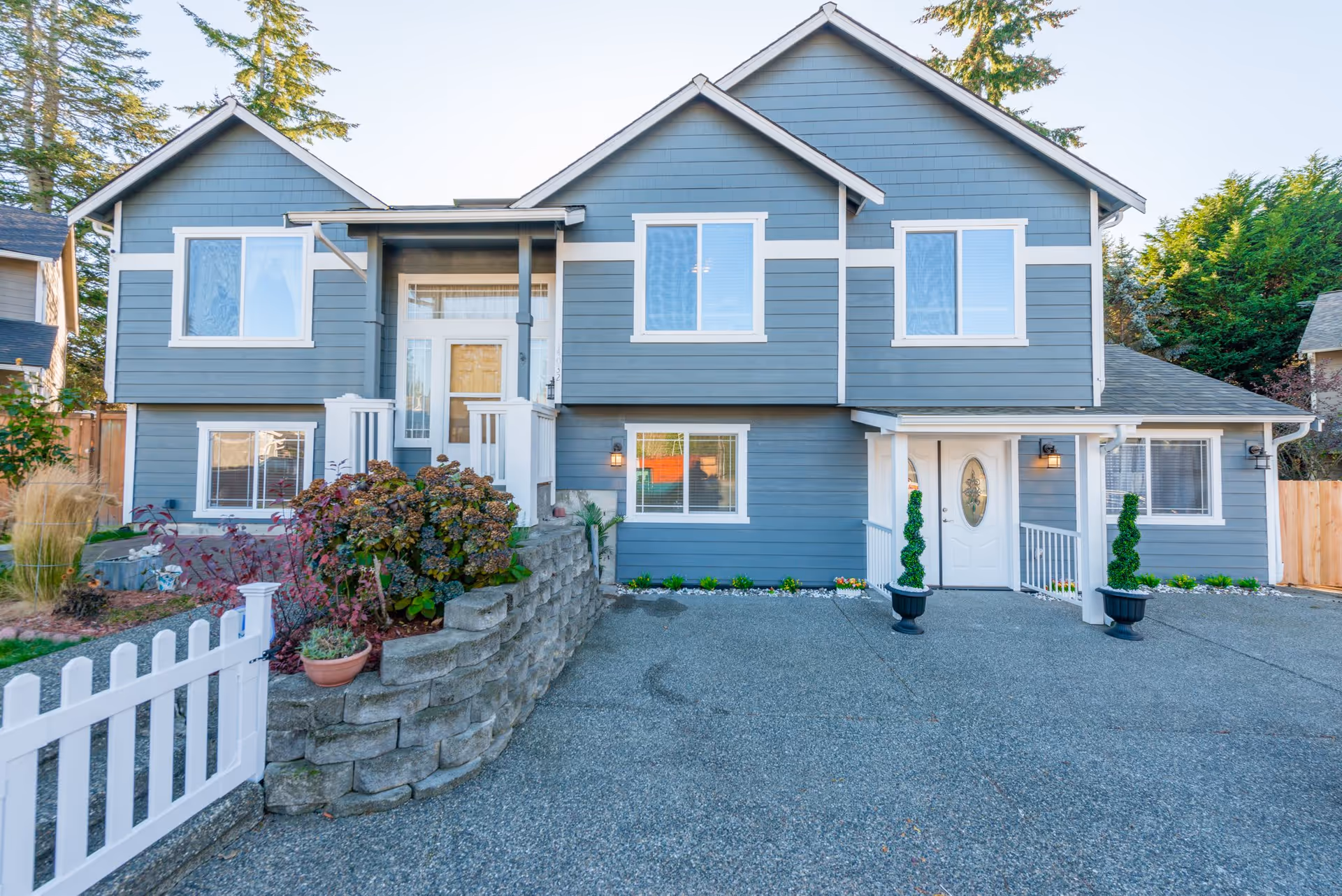 Front exterior view of a two-story gray house with white trim, a white picket fence, and a paved driveway. There are plants and shrubs along the front, and the entrance features a small porch with white railings and two potted topiary plants.
