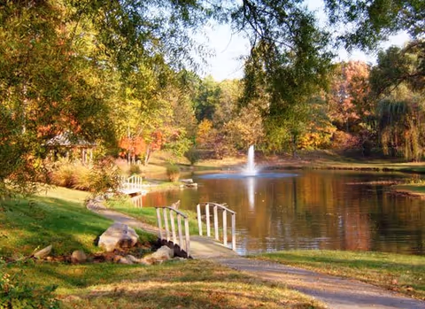 A peaceful outdoor scene featuring a small pond with a water fountain in the center, surrounded by trees with autumn foliage. A curved walking path runs alongside the pond, with a small white footbridge crossing a narrow section of the water. The grass is green and the sky is clear.