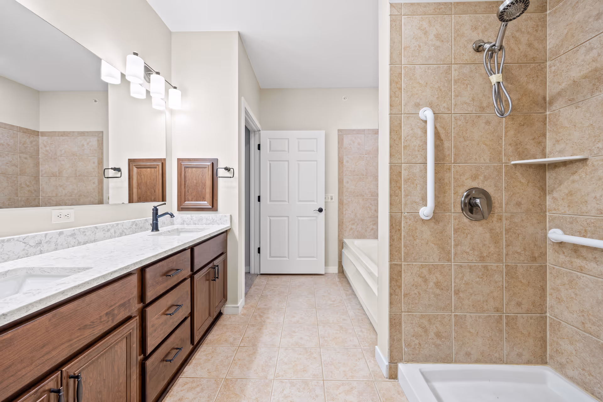 A clean and modern bathroom featuring a large vanity with a marble countertop and dark wood cabinets, a wide mirror with three light fixtures above, a tiled walk-in shower with grab bars and a handheld showerhead, and a bathtub partially visible in the background.