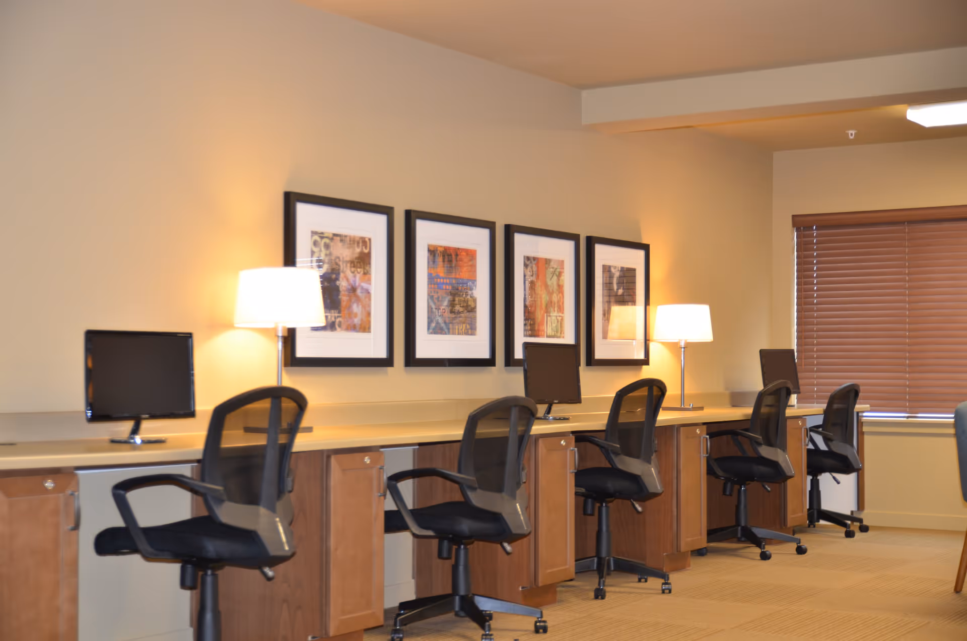 A row of four black office chairs in front of a long wooden desk with three computer monitors and two table lamps. Four framed abstract artworks hang on the beige wall above the desk. A window with closed brown blinds is visible on the right side.