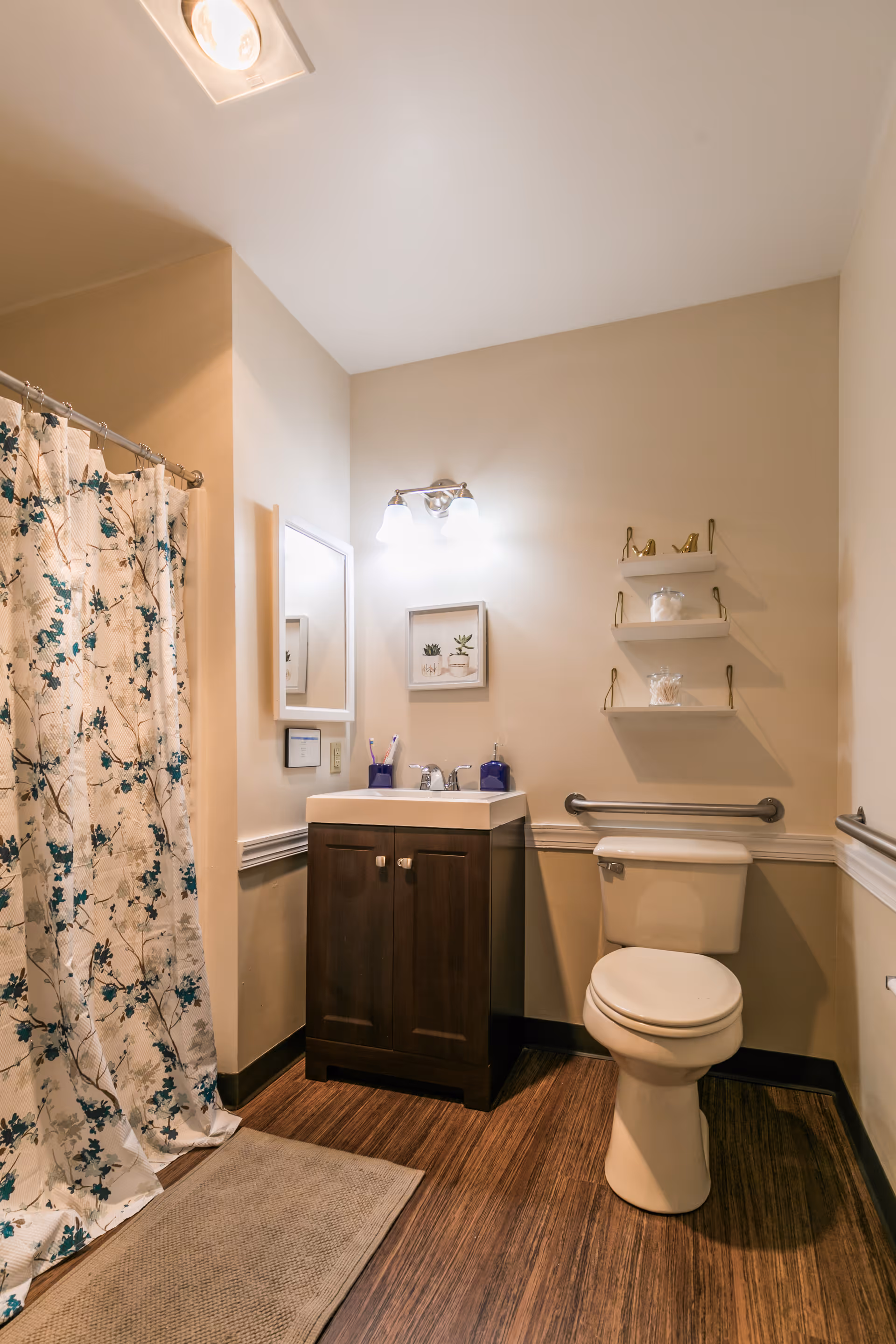 A clean bathroom with a wooden floor, a white toilet, a dark wood vanity with a white sink, a mirror above the sink, a light fixture with two bulbs, a shower with a floral patterned curtain, and three small white shelves on the wall holding decorative items.