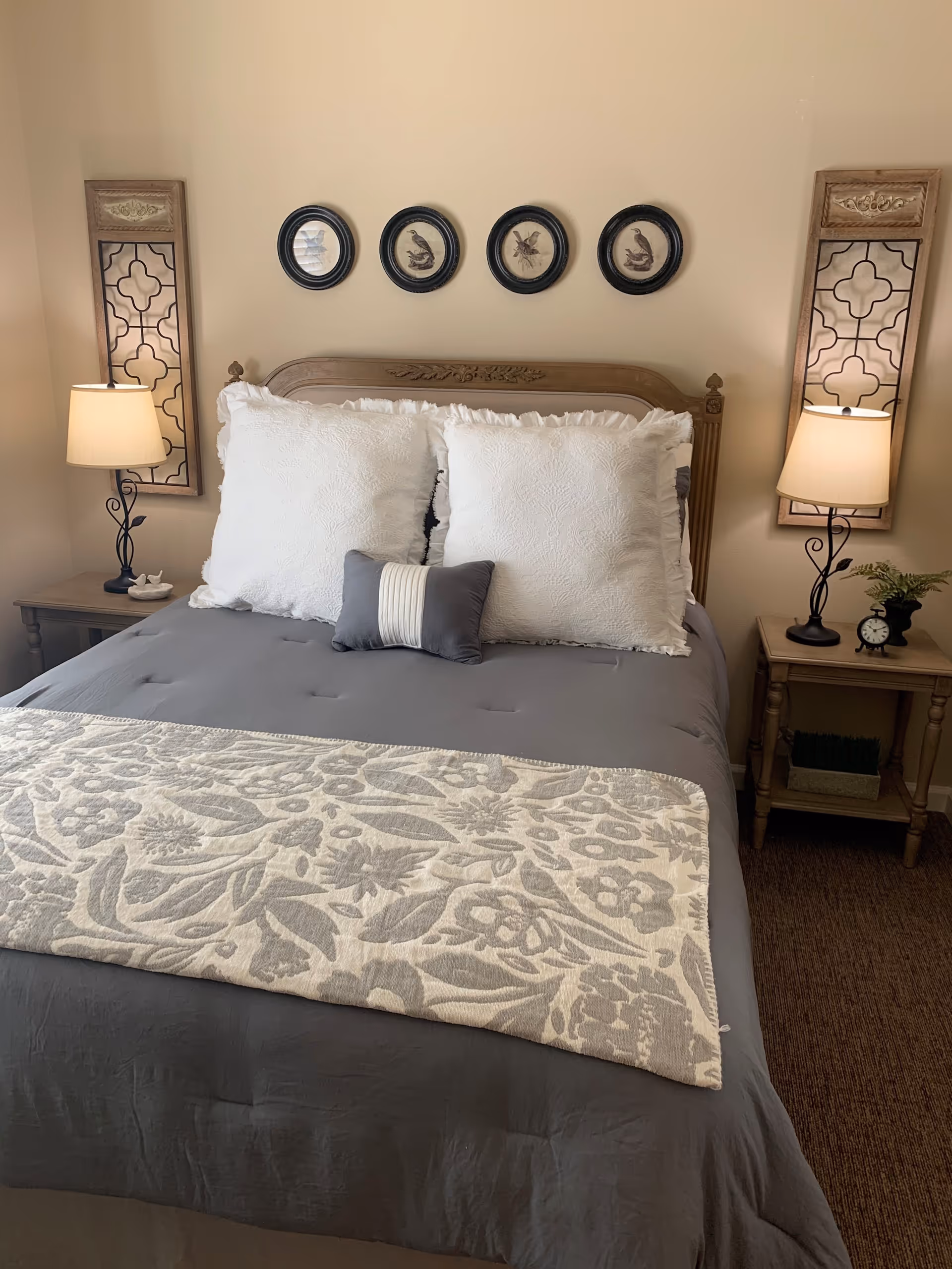 A neatly made bed with a wooden headboard, two large white pillows, and a smaller decorative pillow in front. The bed has a gray comforter and a patterned throw blanket at the foot. On each side of the bed are wooden nightstands with table lamps, decorative mirrors, and small decor items. Four framed circular artworks hang on the wall above the headboard.