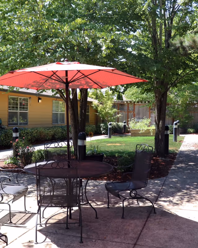Patio courtyard with a red umbrella over a metal table and chairs, surrounded by trees and a single-story building.