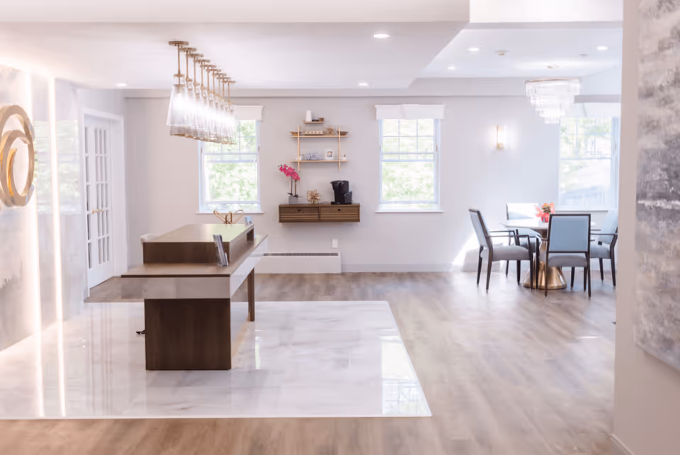 Bright and modern interior space with a wooden reception desk featuring a row of pendant lights above it. The floor is a combination of light wood and white marble tiles. In the background, there are two windows with white trim, a small wall-mounted shelf with decorative items and a coffee maker, and a dining area with a round table and four chairs near a window with a chandelier overhead.