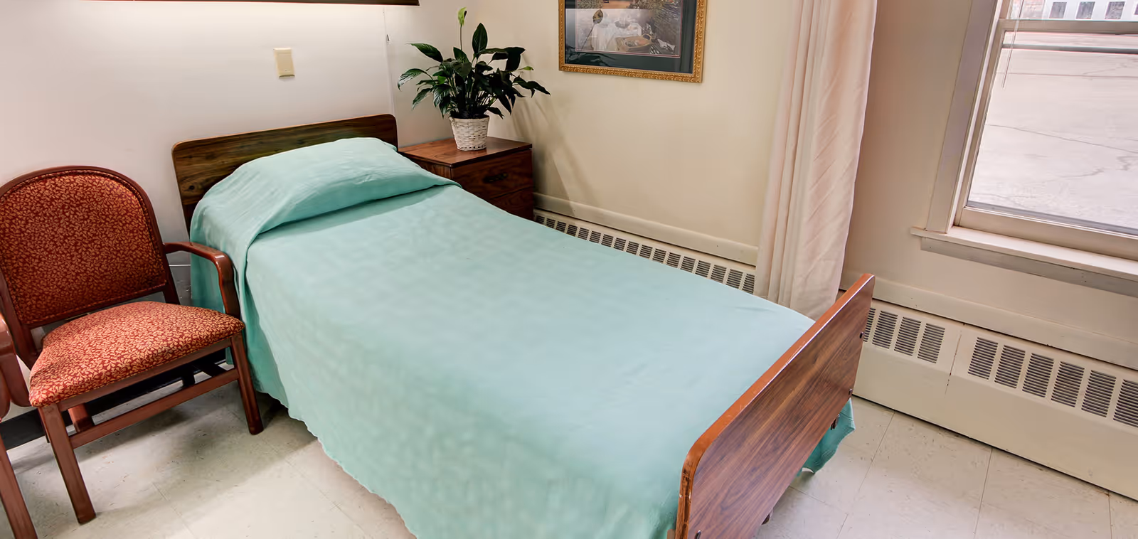 Small bedroom with a single bed covered in a light green bedspread, a wooden nightstand with a plant, an upholstered chair, and a window.