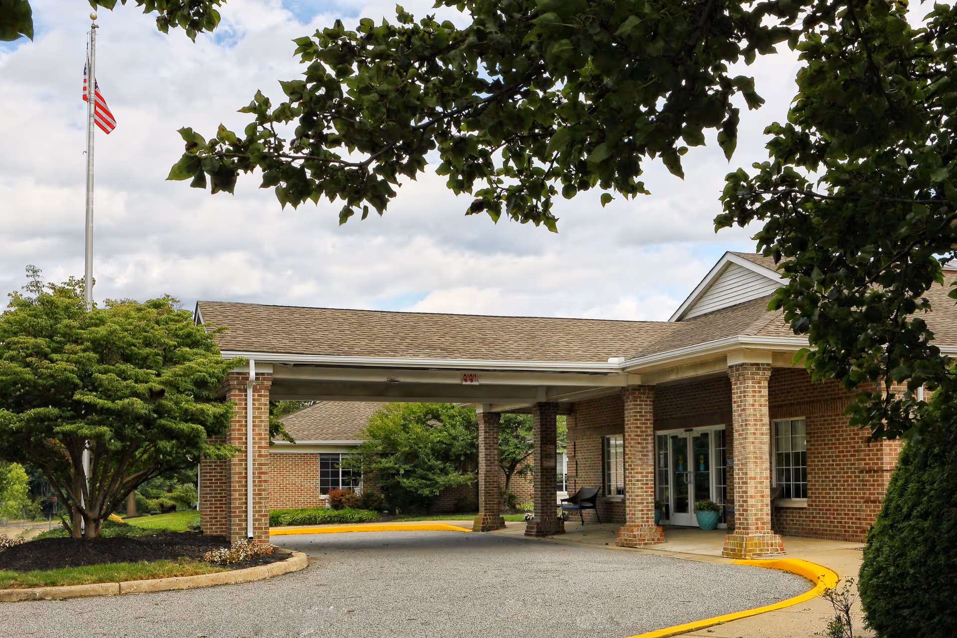 Entrance of a brick building with a covered driveway, surrounded by trees and greenery, with an American flag on a flagpole to the left.
