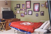 A bedroom with a hospital-style bed covered with a red blanket featuring a white logo. The bed is positioned against a green wall decorated with multiple framed photos and pictures. There is a wooden dresser with a television on top and a small folding table next to it.