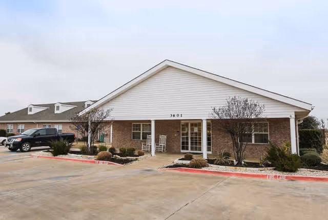 Exterior view of a single-story brick building with a white triangular roof and the number 3601 above the entrance. There are two white rocking chairs on the front porch, small trees, and shrubs in landscaped beds. A black SUV is parked on the left side in a concrete parking area.