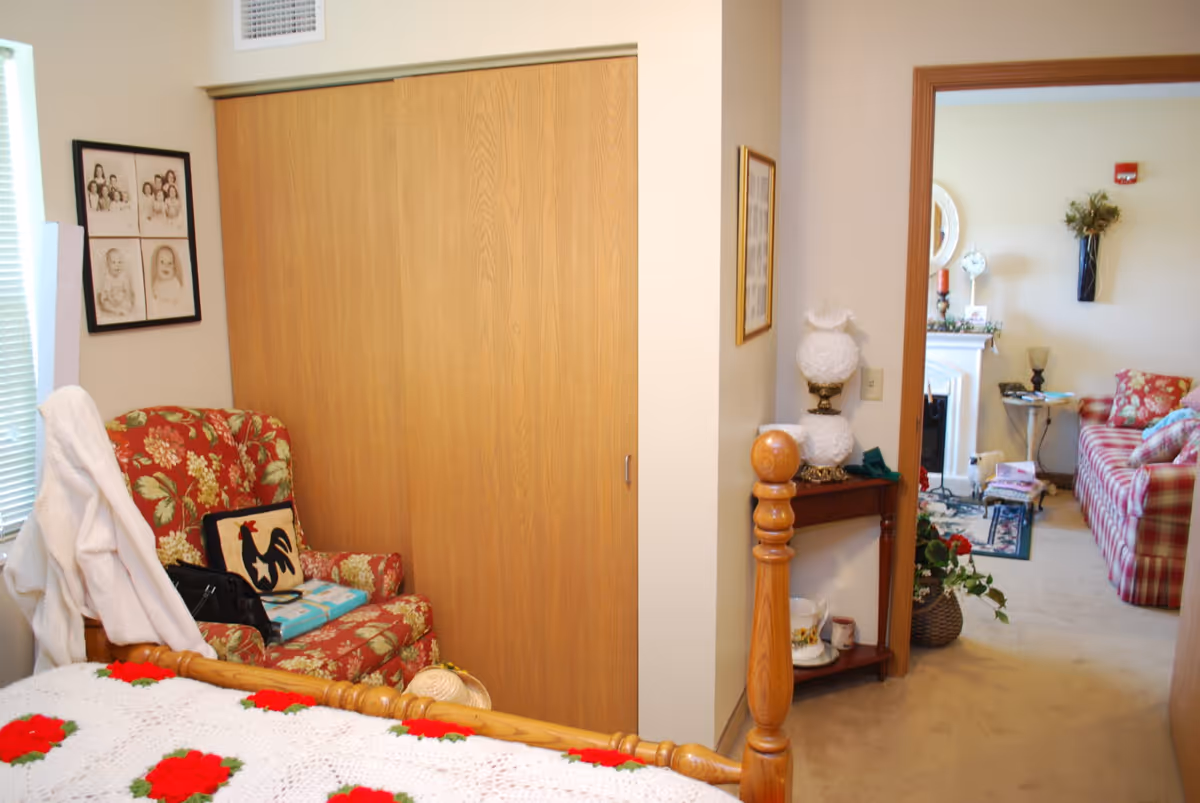 Small bedroom featuring a wooden bed with a crocheted blanket, a floral armchair next to sliding closet doors, and a view into a furnished living area.