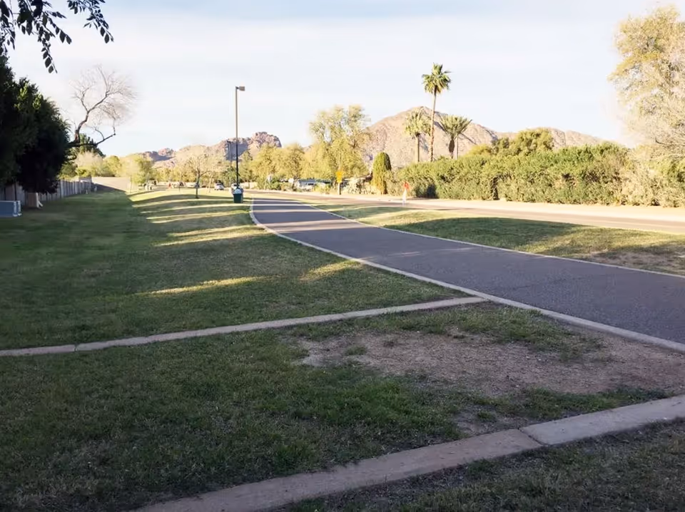 A paved walking path runs through a grassy area with trees and bushes on either side. In the background, there are mountains and a clear sky. Palm trees and a street lamp are visible along the path.