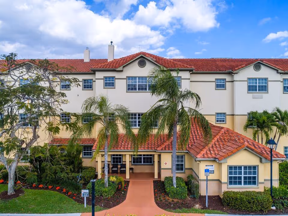Exterior view of Sonata Coconut Creek facility showing a three-story building with beige and light yellow walls, red tiled roof, multiple windows, palm trees, and a landscaped garden with flowers and shrubs under a partly cloudy sky.
