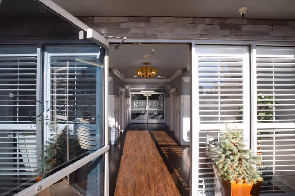 Entrance hallway of Redding Post Acute facility with glass doors at the far end displaying the facility name. The hallway has wooden flooring, white wainscoting on the walls, a gold chandelier on the ceiling, and large windows with white blinds on the right side. A small potted plant is visible near the entrance.