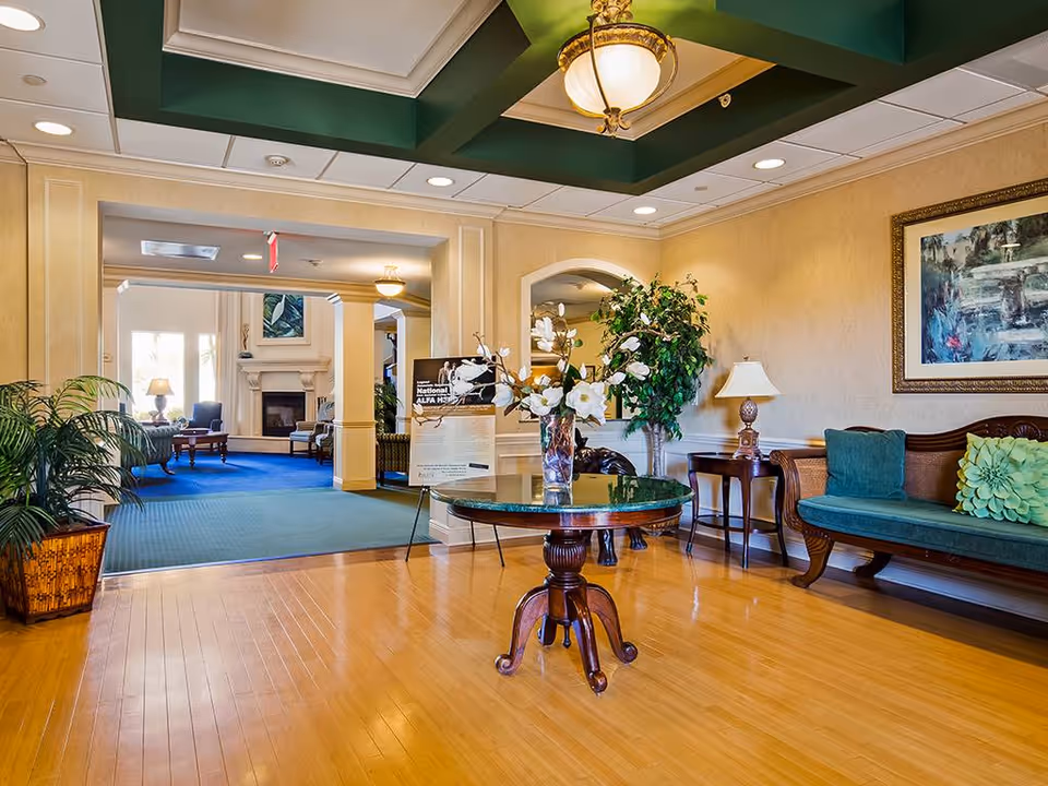Bright senior living lobby with a round table topped with flowers, seating benches, plants, and a view into a sitting room with a fireplace.