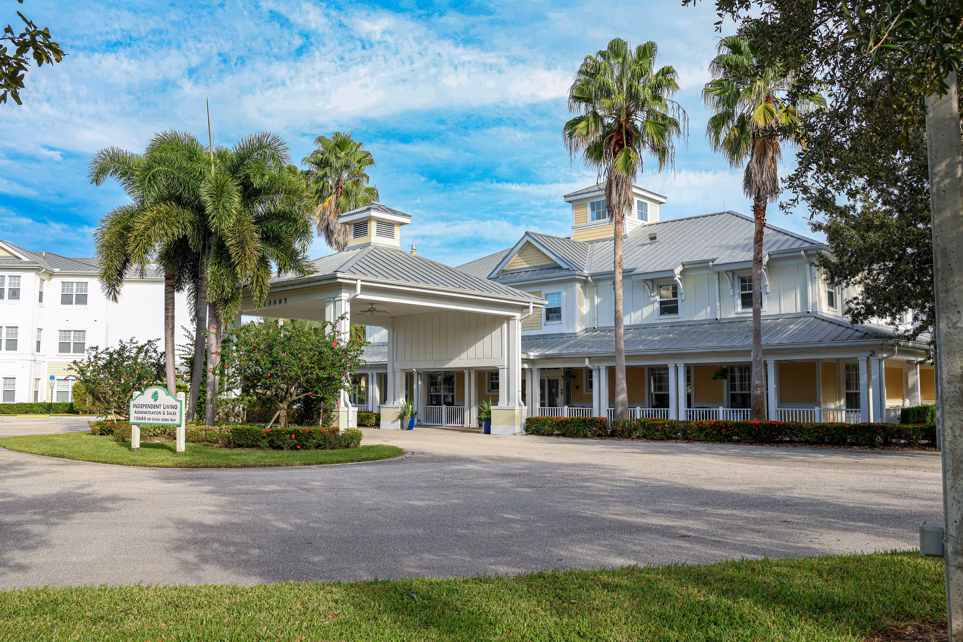 Exterior view of a senior living facility building with a covered entrance, palm trees, and a sign indicating independent living apartments and sales. The building has a light yellow and white color scheme with a metal roof under a partly cloudy blue sky.