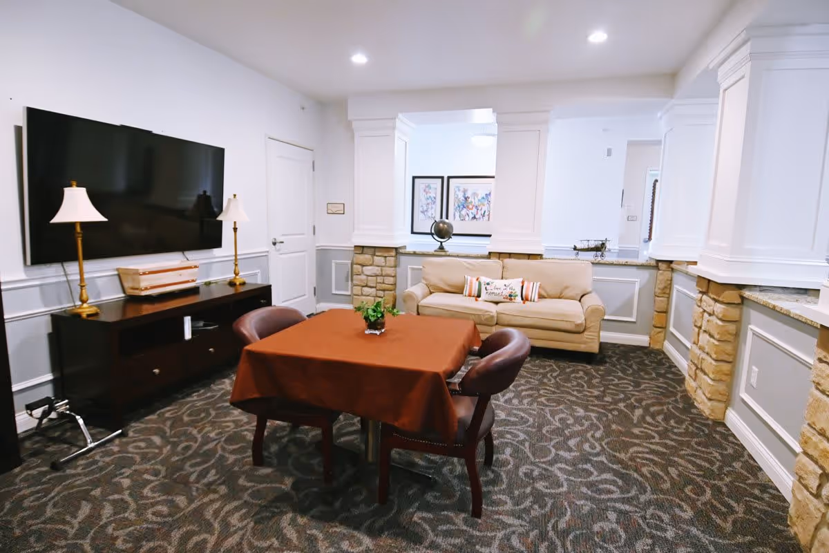 A cozy living room area with a beige sofa adorned with decorative pillows, a square table covered with a brown tablecloth surrounded by four chairs, a large flat-screen TV mounted on the wall above a dark wooden cabinet with two lamps, and decorative stone pillars with white paneling.