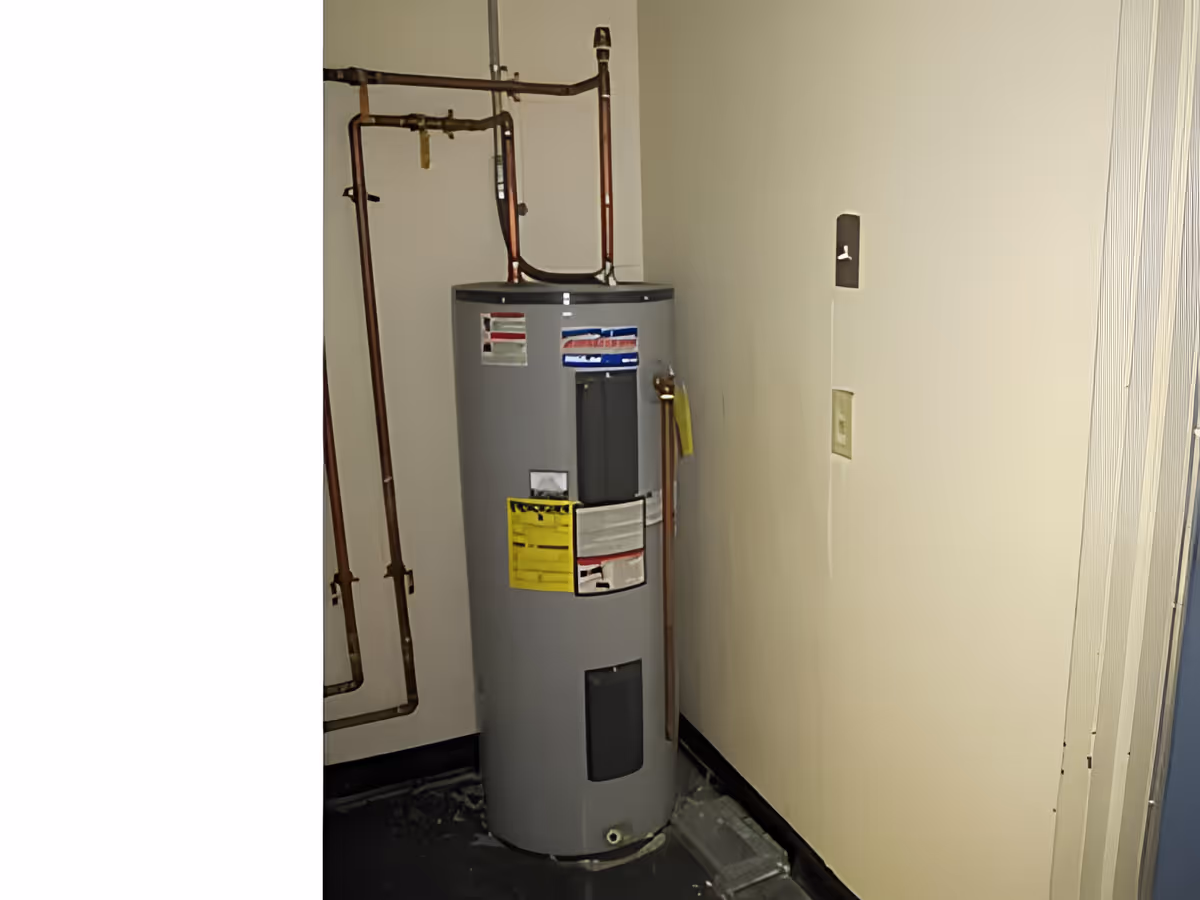 A gray water heater tank installed in a corner of a room with copper pipes connected to it. The walls are plain and light-colored, and there is a small electrical outlet on the right wall.