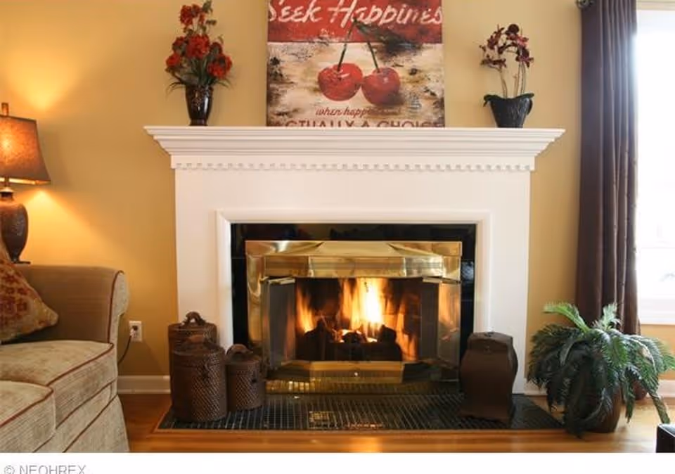 A cozy living room corner featuring a lit fireplace with a white mantel. On the mantel are two vases with red flowers and a framed artwork with cherries and the words 'Seek Happiness'. To the left is a beige sofa with a patterned pillow and a lit table lamp. To the right of the fireplace is a green potted plant near a window with brown curtains.