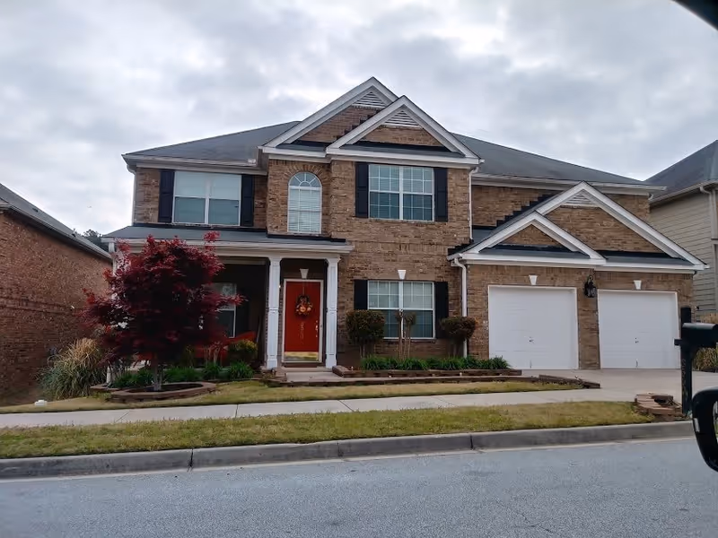 Front exterior view of a two-story brick house with a red front door decorated with a wreath, two white garage doors, and a small landscaped yard with a tree and shrubs.