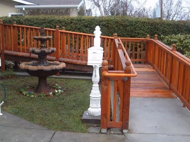 Outdoor area with a wooden ramp and railing leading up to a building entrance. There is a three-tiered stone fountain on a grassy patch next to the ramp, and a white decorative mailbox or post in front of the ramp. Trees and bushes are visible in the background.