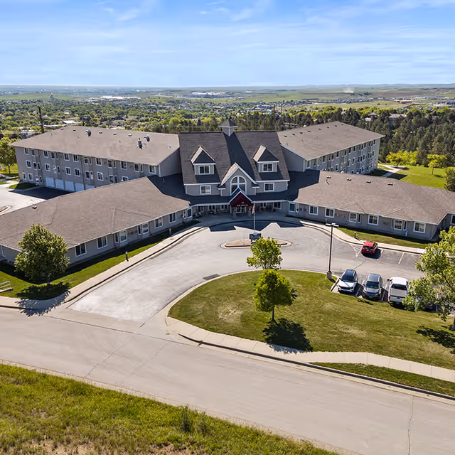 Aerial view of Good Samaritan Society - Echo Ridge - The Manor, a large senior living facility with a central building and two wings extending on either side. The building has a peaked roof and multiple windows. There is a circular driveway with a small landscaped island in front, several parked cars, and green lawns surrounding the facility. The background shows a wide landscape with trees and distant hills under a clear blue sky.
