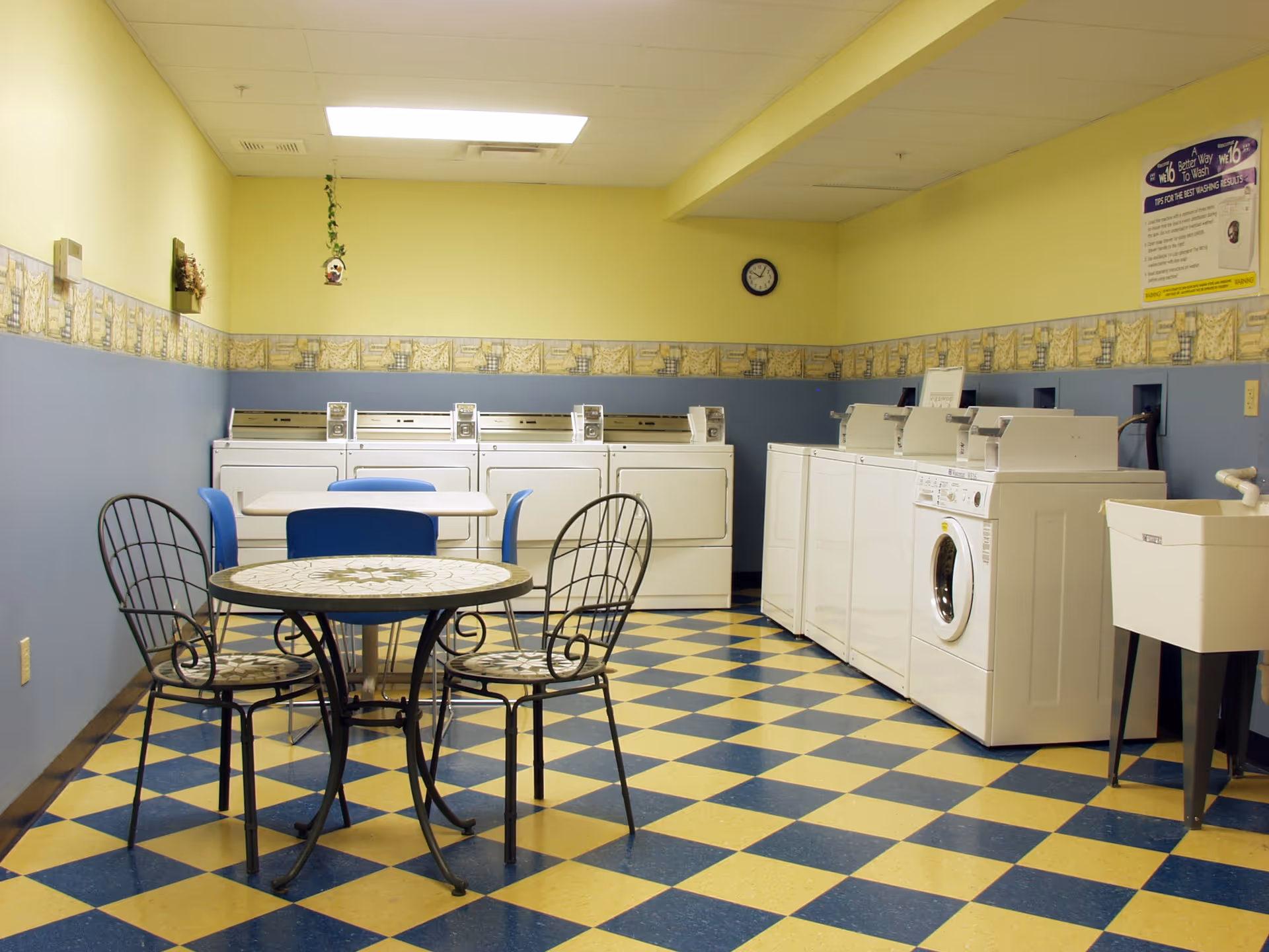 A laundry room with several white washing machines and dryers along the walls. There are two tables with chairs in the center of the room. The floor has a blue and yellow checkered pattern, and the walls are painted yellow and blue with a decorative border. A clock and a handwashing instruction poster are visible on the walls.