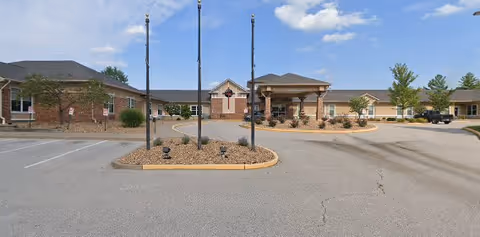 Front exterior view of Arbor Hills Nursing and Rehabilitation Center showing a single-story building with a covered entrance, surrounded by a parking lot and landscaped areas with small trees and shrubs under a blue sky with some clouds.