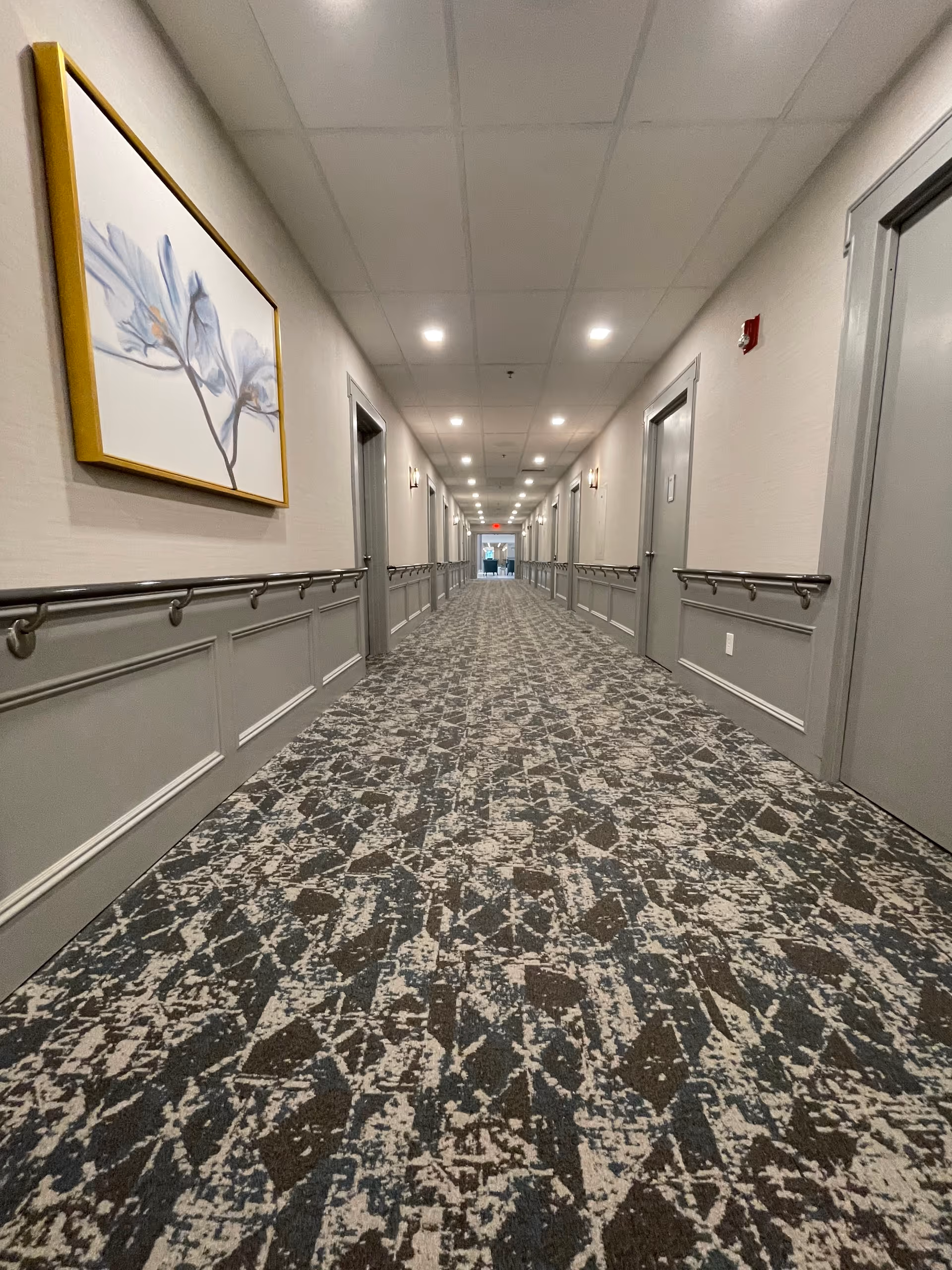 Long indoor hallway in a senior living facility with patterned carpet, gray doors on both sides, handrails along the walls, ceiling lights, and a framed floral artwork on the left wall.