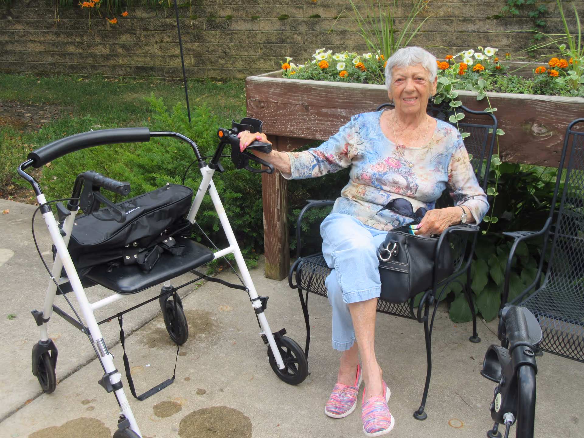 An elderly woman with short white hair sitting on a black metal chair outdoors next to a raised wooden planter box filled with flowers. She is wearing a floral long-sleeve shirt, light blue pants, and pink slip-on shoes. A white walker with a black seat and bag is positioned nearby on the concrete ground.