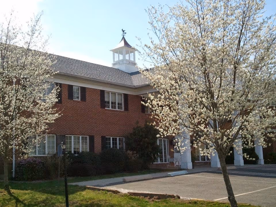 Exterior view of a two-story brick building with white-trimmed windows and a cupola on the roof, surrounded by blooming trees with white flowers and a parking area in front.