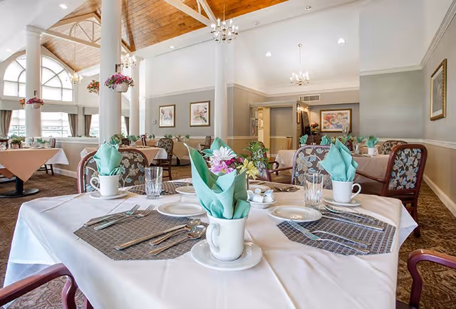 A bright and spacious dining room in a senior living facility with tables set for a meal. Each table has white tablecloths, placemats, cups with green folded napkins, plates, cutlery, and small flower arrangements. The room features high ceilings with wooden panels, chandeliers, large arched windows, and framed artwork on the walls.