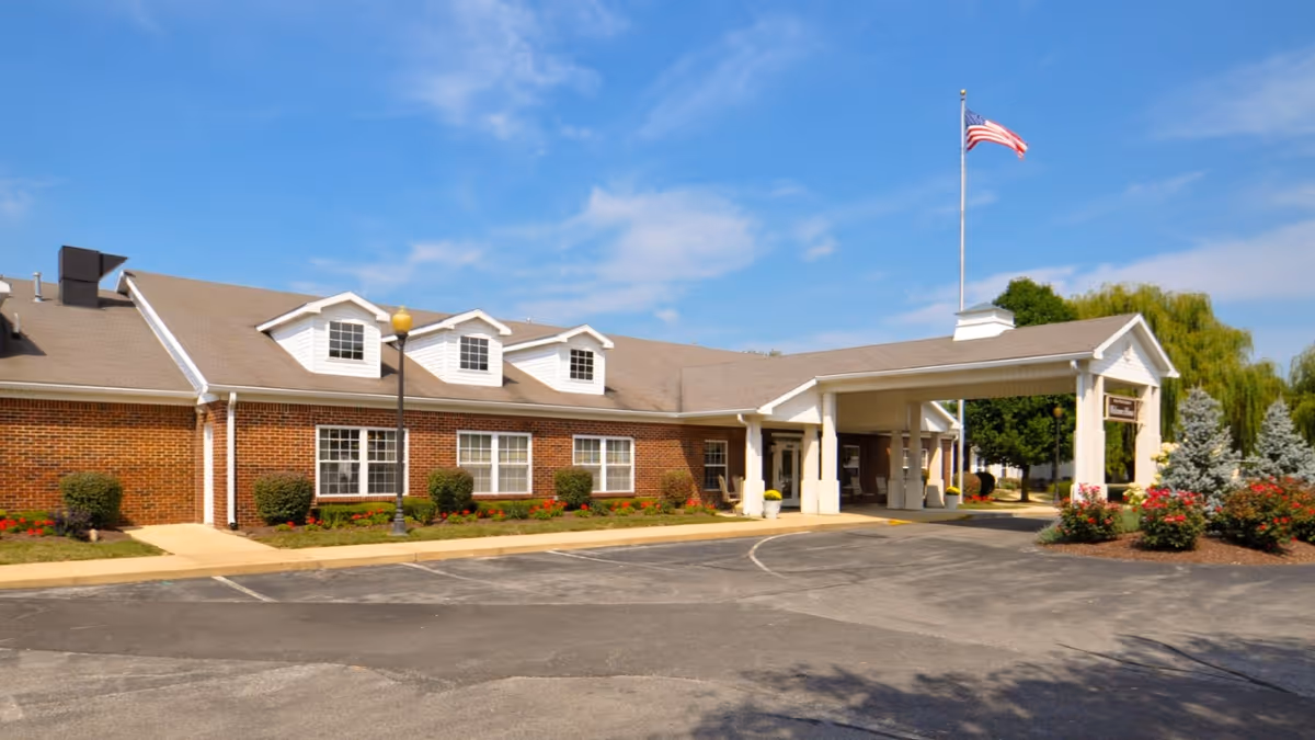 Exterior view of a single-story brick building with a covered entrance, surrounded by landscaped bushes and flowers, under a blue sky with some clouds. An American flag is flying on a flagpole near the entrance.