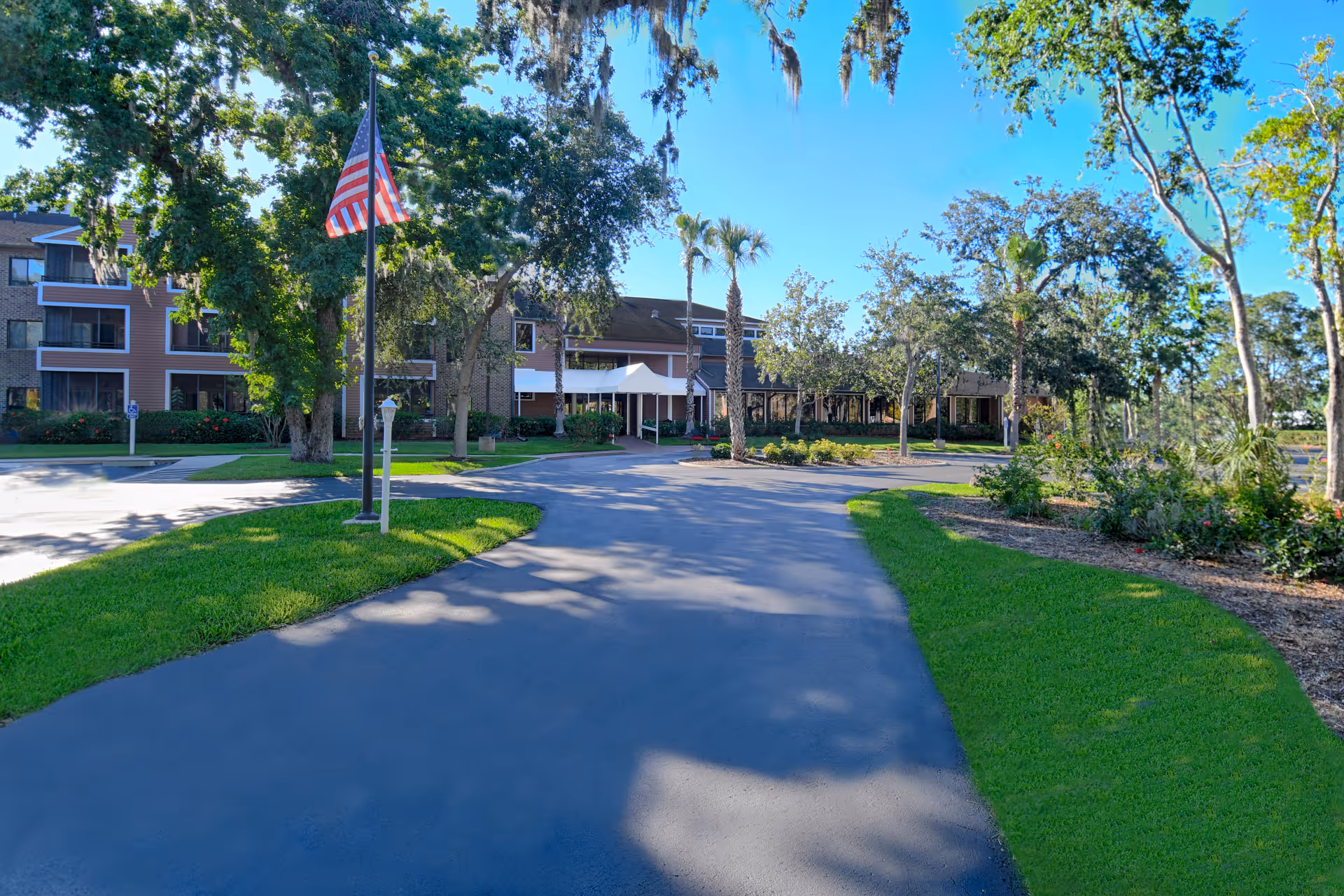 View of the exterior entrance of a senior living facility with a paved driveway, green lawns, trees including palm trees, and an American flag on a flagpole. The building has multiple floors with balconies and large windows under a clear blue sky.