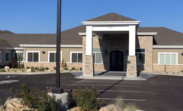 Front exterior view of a single-story assisted living facility building with a covered entrance supported by stone pillars, surrounded by a parking lot and landscaping with bushes and rocks under a clear blue sky.