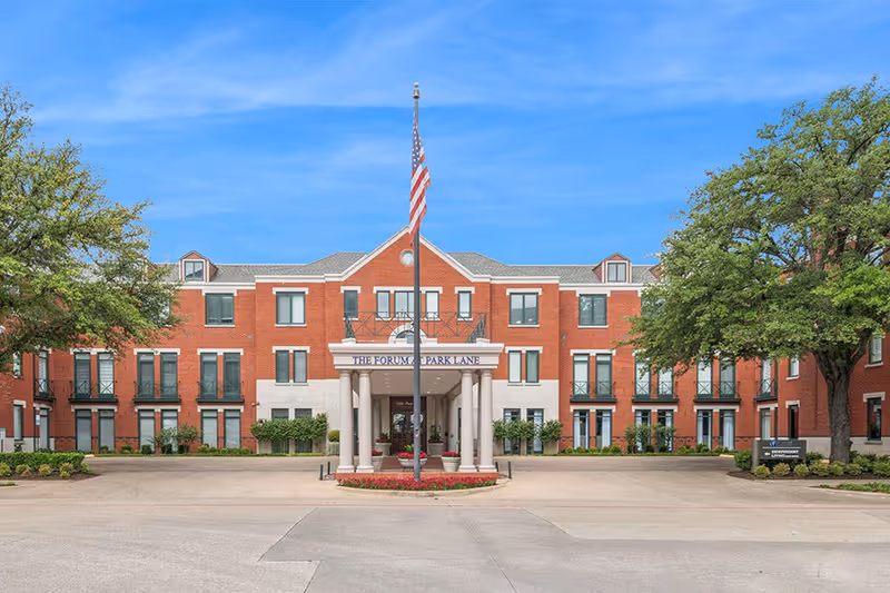 Front exterior view of The Forum at Park Lane, a three-story red brick building with white stone accents, multiple windows, and a covered entrance supported by columns. An American flag is flying on a flagpole in front of the entrance, with trees and landscaping on either side.
