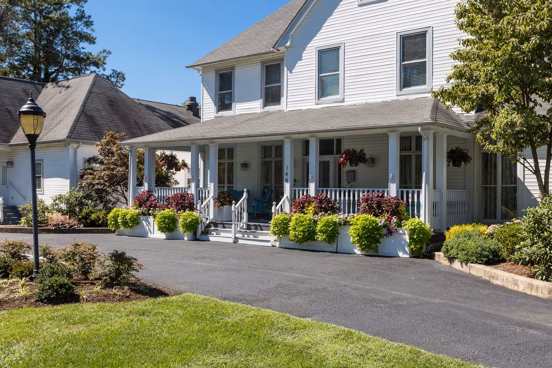 Exterior view of a white two-story house with a covered porch decorated with hanging flower baskets and planter boxes filled with green and purple plants. The house has multiple windows and a paved driveway in front. There is a street lamp and well-maintained landscaping with bushes and grass surrounding the house.