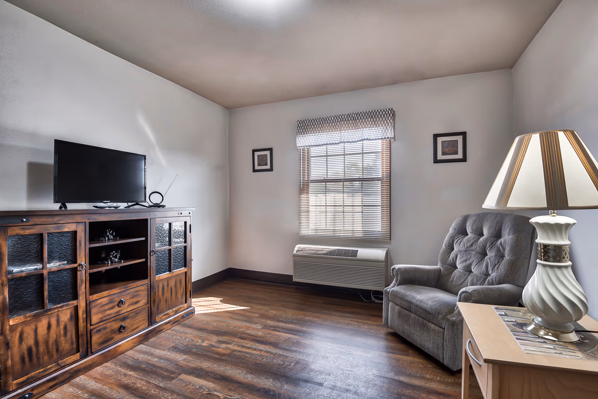 Small living room with a TV on a wooden cabinet, an upholstered armchair, a side table with a lamp, and a window with blinds.