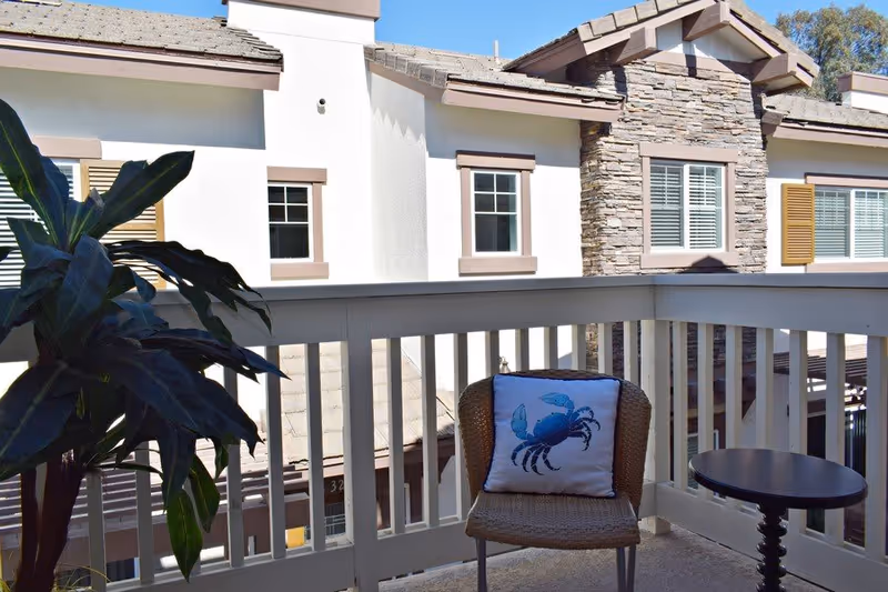 A small balcony with a wicker chair featuring a blue crab design pillow, a round black side table, and a large leafy plant. The balcony overlooks neighboring buildings with white walls, stone accents, and windows with shutters.