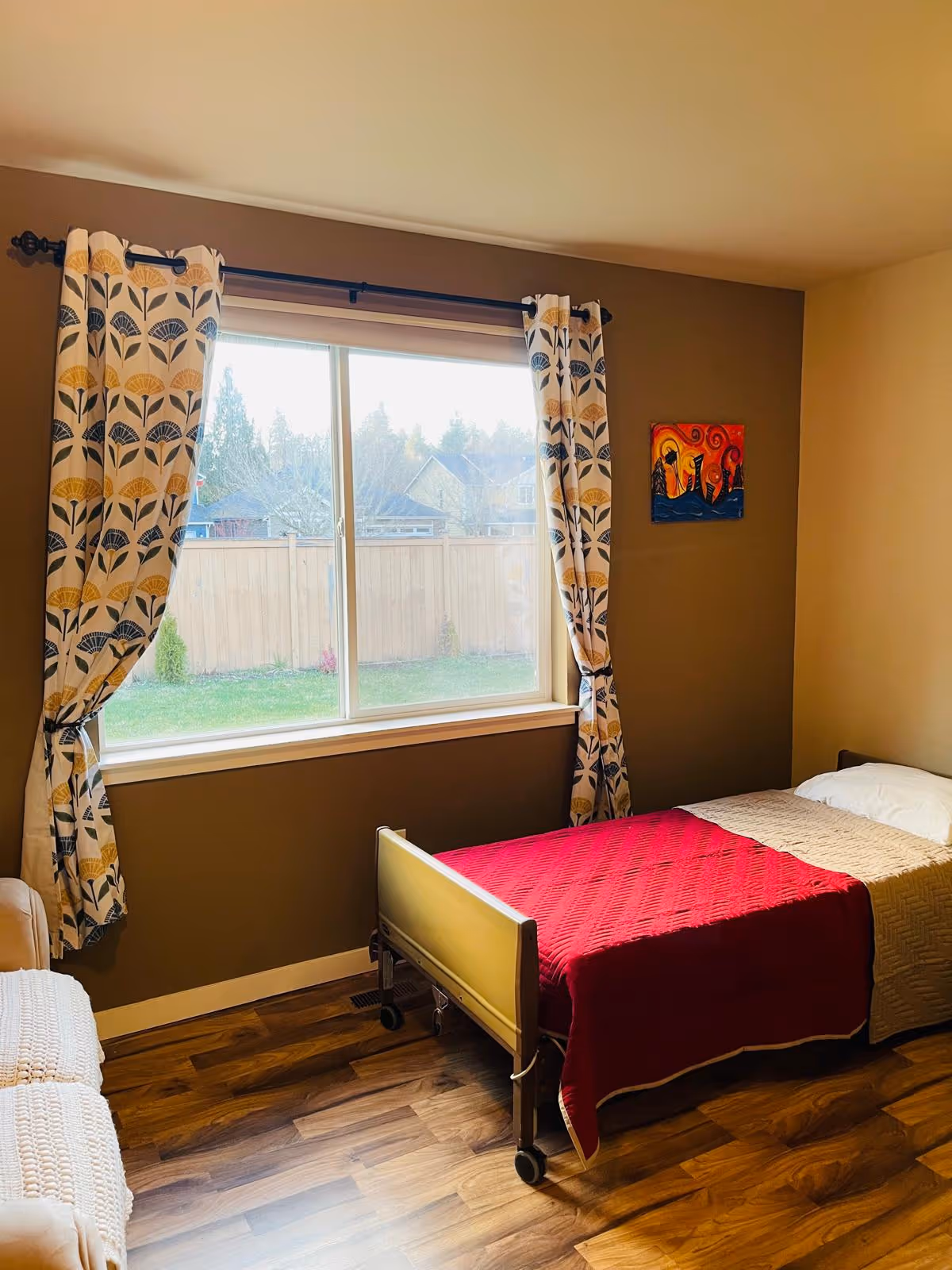 A small bedroom with a single bed covered with a red blanket and beige quilt. The room has wooden flooring, a large window with patterned curtains, and a small colorful painting on the wall. There is also a beige armchair partially visible on the left side.